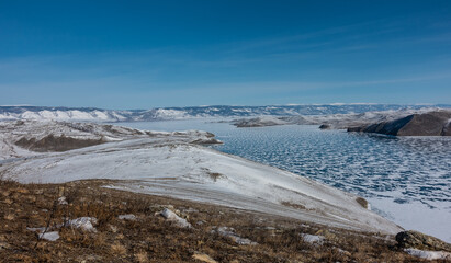 Panorama of the frozen lake. The icy surface is covered with snow, like lace. Around are snow-capped hills. Dry grass in the foreground. Sunny winter day, blue sky. Baikal