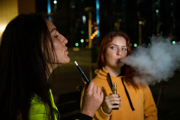 Two caucasian women smoke vape and hookah outdoors at night.