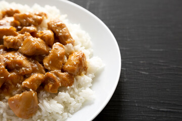 Homemade Orange Chicken with White Rice on a white plate on a black surface, side view. Copy space.