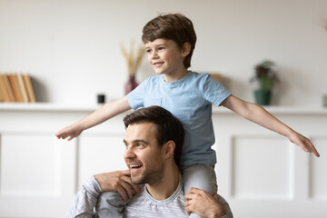 Excited small Caucasian 7s boy child sit on young father shoulder back have fun at home on family free weekend together. Happy little son kid play with dad, engaged in funny game activity.