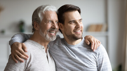 Smiling young Caucasian man and older father hug look in window distance dreaming imagining together. Optimistic mature dad and adult grownup son embrace visualize happy future. Vision concept.
