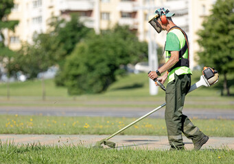 Municipal gardener landscaper man worker cutting grass with string trimmer machine along city street