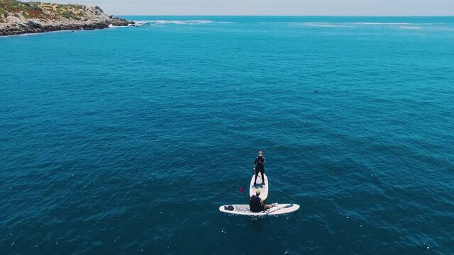 Aerial drone shot over the beach, cinematic and static stabilized video of two unrecognizable people learning stand up paddle boarding