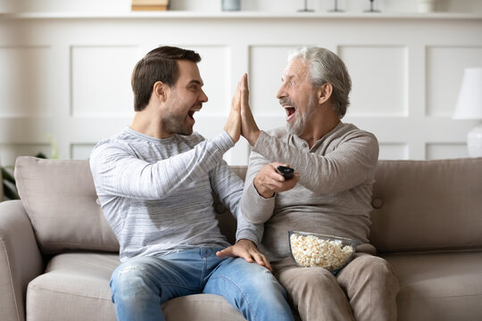 Happy older Caucasian father and adult grownup son give high five celebrate win watching TV together. Overjoyed millennial man have fun enjoy at home with senior dad with football game.