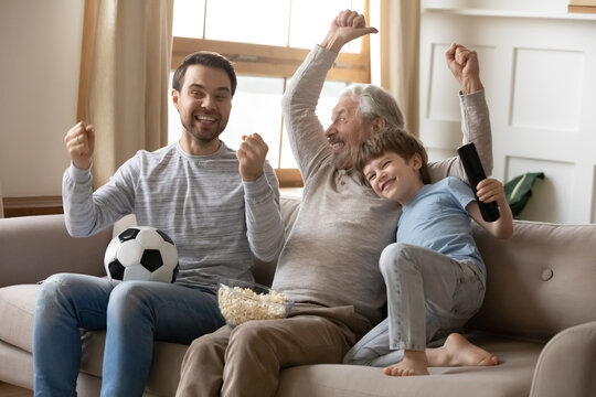 Smiling Three Generations Of Caucasian Men Have Fun Cheering Watching Football On TV With Popcorn Snacks. Overjoyed Boy Child Enjoy Television With Young Father And Mature Grandfather. Fan Concept.