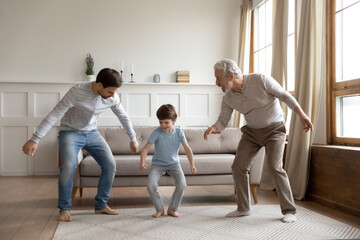 Happy three generations of Caucasian men have fun dancing together in cozy living room on weekend....