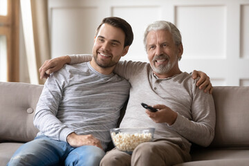 Smiling senior 60s Caucasian father and adult son sit relax on sofa at home watching TV on weekend together. Happy older dad and grownup millennial male child rest enjoy program on television.