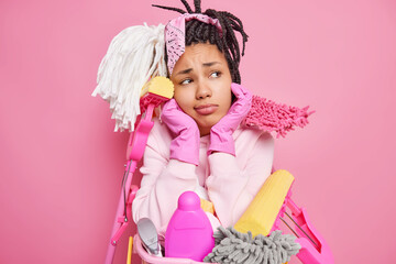 Housekeeping concept. Sad bored woman with dreadlocks holds chin looks sadly at camera surrounded by cleaning supplies holds near basin of laundry isolated over pink background. Cleanup day.