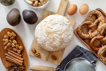 Fresh bread on wooden board, avocados, eggs, pretzel, nuts and spices on white table. Ingredients for delicious toasts for breakfast. Top view, flat lay.