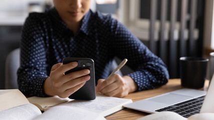 Close up Indian woman holding phone, writing notes, watching webinar, writing down notes, sitting at table with books and laptop, student preparing for exams at home, businesswoman planning workday