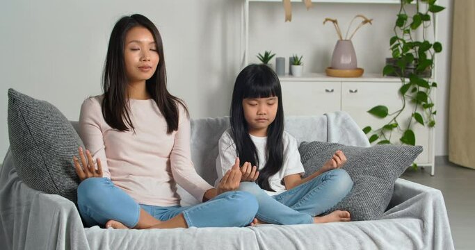 Asian Buddhist family traditions, ethnic Korean mother teaches her little beloved daughter to meditate keep calm two people sit on sofa in lotus position in asana with closed eyes, calm mind and soul