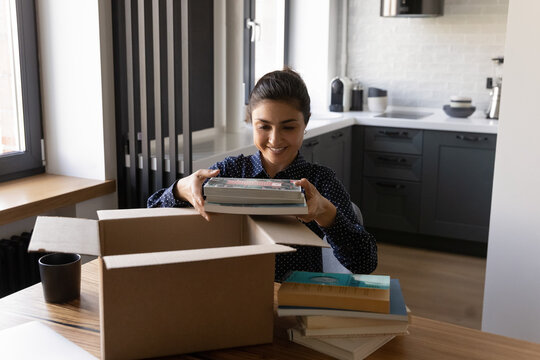 Smiling Indian woman unpacking parcel, holding books, sitting at table, satisfied customer received online store order, delivery service, happy female packing belongings, preparing for relocation