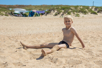 A blond boy sits on the sand by the sea and smiles. A tent camp in the background. Summer happy vacation. Sea holidays.