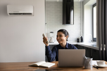 Smiling Indian businesswoman or student using air conditioner remote controller, working online at...