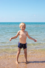 Little blond boy in swimming trunks is playing on the seashore with sand and water, smiling, standing in the water. Summer happy vacation.