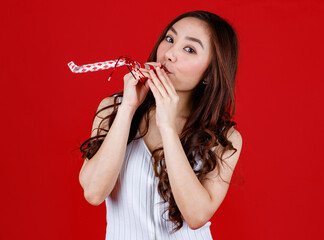 Funny young and cure Asian girl playing and blowing party whistle. Studio shot on red background