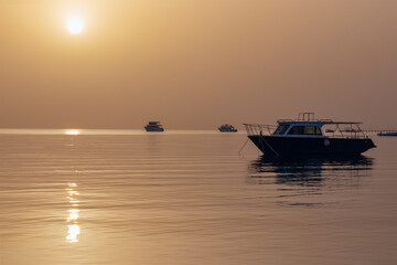 Naklejka premium Dawn on the Red Sea in Egypt, Makadi Bay, calm sea, silhouettes of boats, golden disc of the sun on the horizon, reflection of sunlight on the water surface
