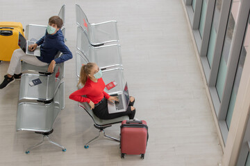 Fototapeta na wymiar Two Caucasian couple tourists wearing hygiene protective mask on faces sitting together on chair in airport with social distancing sign. Concept for new normal traveler during coronavirus outbreak