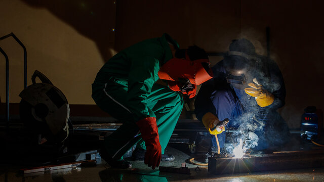 Mechanics Workers Work Overtime Hardly At Night In A Factory. Engineers Wearing Safety Outfits With Mechanic Jumpsuits, Gloves, Boots, And Welding Helmets Working On Metal Welding