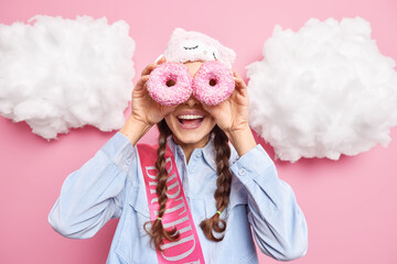 Positive young woman smiles gladfully covers eyes with glazed sweet doughnuts enjoys eating delicious bakery wears sleepmask shirt and birthday ribbon isolated over pink background clouds above