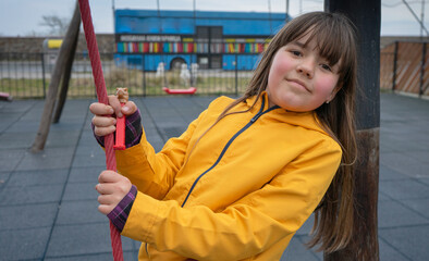 A child on the playground, a girl with long brown hair is played and smiles in a yellow jacket