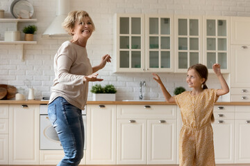 Overjoyed mature grandmother and cute little granddaughter dance in kitchen together on weekend at...