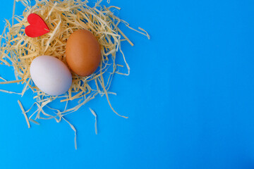 A brown and white chicken egg and a small red wooden heart lie on hay on a blue background. Easter card concept