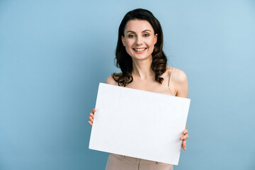 Beautiful brunette girl with holds a white sheet of paper on a blue background. Place for text, banner, surface for inscriptions and advertising.