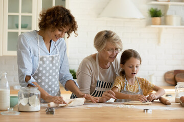 Happy three generations of Caucasian women have fun cooking together bread or pie in home kitchen. Cute little 9s girl child baking with mature 60s grandmother and young mother, prepare dessert. - obrazy, fototapety, plakaty