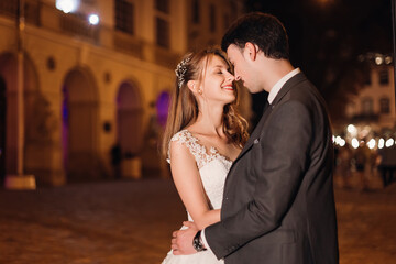 Gentle and loving newlyweds hug against the backdrop of the night city and light bulbs. Stylish groom and beautiful bride