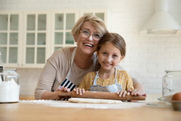 Portrait of smiling elderly 60s Caucasian grandmother and little 8s granddaughter baking together in home kitchen. Happy mature 50s granny and small grandchild cooking pie or cookies on weekend.