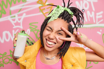 Cheerful teen girl with dreadlocks golden teeth makes peace or victory gesture makes graffiti with aerosol spray dressed in fashionable clothes. Millennial generation and street art concept. - obrazy, fototapety, plakaty