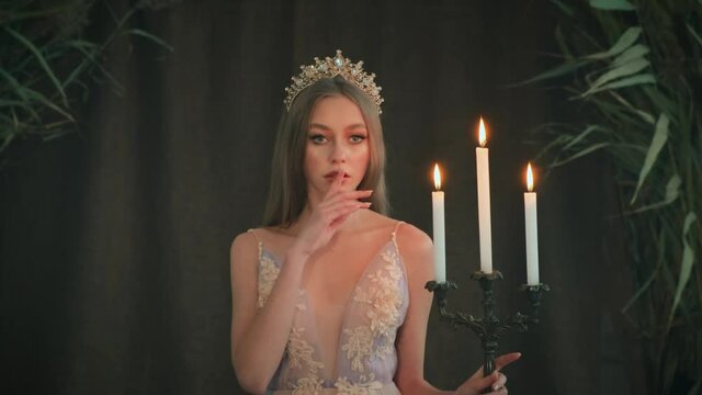 girl princess stands in room. Woman queen is holding candlestick burning candles in hand. Pink dress, long loose blonde hair gold royal crown. finger near lips, silence gesture order not to make noise