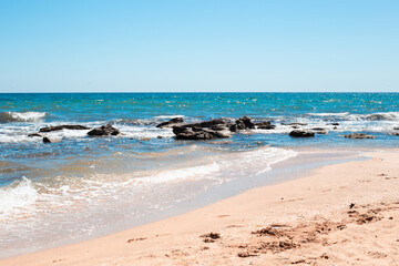 Seashore with blue water beating against stones and a sandy beach. Sea sunny summer landscape