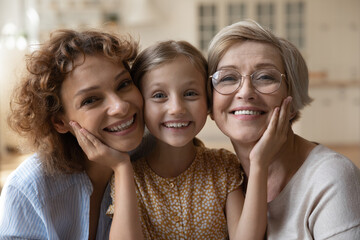 Close up headshot portrait of smiling three generations of Caucasian women pose together relax at home. Happy little girl child with young mother and senior grandmother embrace show love care.