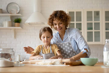 Smiling young Caucasian mother and little 8s daughter roll dough cook pie or pastry together in...