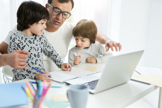 Cute little latin boy spending time with his father and brother at home. Businessman using laptop while working from home during lockdown and watching children