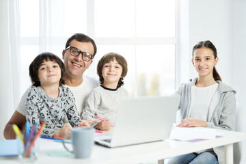Happy latin father and kids smiling at camera, sitting together at the table at home. Man using...
