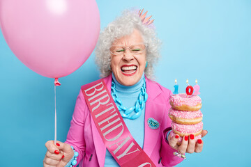 Photo of positive elderly woman celebrates 102nd birthday holds delicious doughnuts and inflated ballon wears festive clothes waits for guests accepts congratulations isolated over blue wall