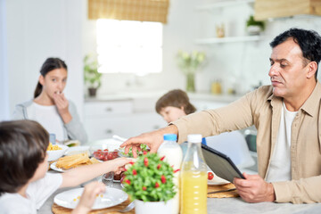 Middle aged father enjoying meal together with his family, using digital tablet while sitting at the table in kitchen at home