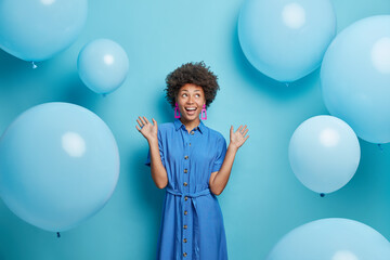 Indoor shot of positive curly haired Afro American woman raises palms dressed in festive dress looks above isolated over blue background with inflated balloons around enjoys birthday celebration