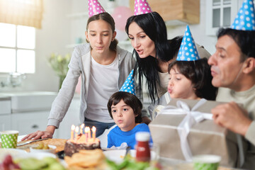 Cheerful latin american family wearing birthday caps, blowing candles on a cake while celebrating birthday together at home