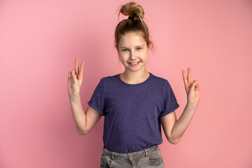 Cheerful child gestures, shows a gesture of peace, victory on the background of a pink wall. Teen girl on a copy space background.