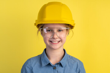 Cheerful, smiling child in a helmet and construction glasses on a yellow background. Girl isolated on blank wall background.
