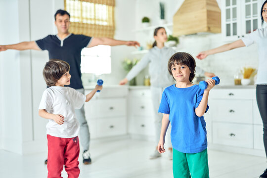 Fitter, healthier, happier. Cute little latin boy smiling at camera, exercising with dumbbell while working out together with his parents and siblings at home