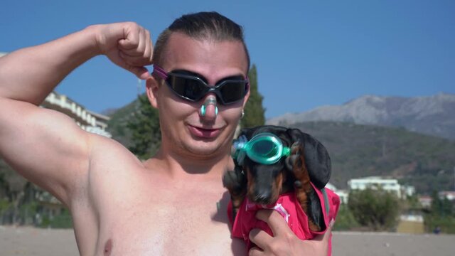 Young athletic man wearing scuba goggles and nose plug for swimming holds funny dachshund puppy in diving gear and flaunts himself in front of camera before going into water, front view.