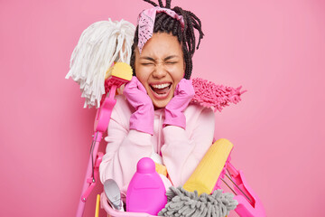 Happy overjoyed housewife expresses positive emotions surrounded with cleaning tools tidies up tools wears rubber gloves isolated over pink background uses different detergents. Housekeeping concept
