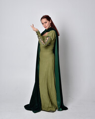 full length portrait of red haired girl wearing celtic, green medieval gown with fantasy velvet cloak. Standing pose isolated against a studio background.