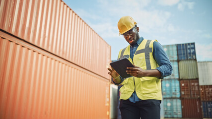 Smiling Portrait of a Handsome African American Black Industrial Engineer in Yellow Hard Hat and Safety Vest Working on Tablet Computer. Inspector or Safety Supervisor in Container Terminal.