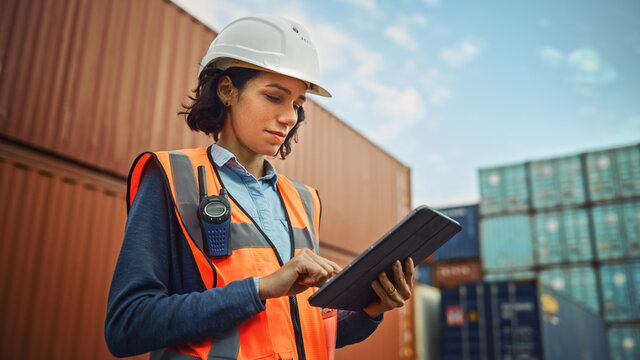 Smiling Portrait Of A Beautiful Latin Female Industrial Engineer In White Hard Hat, High-Visibility Vest Working On Tablet Computer. Inspector Or Safety Supervisor In Container Terminal.